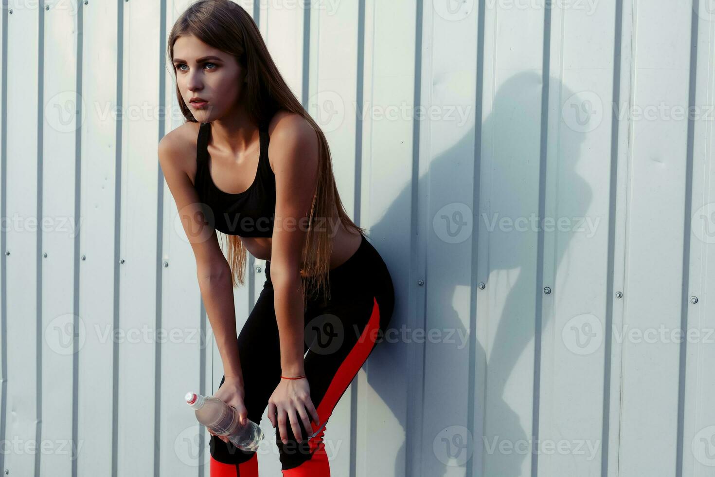 Female athlete drinking water while standing by a grey wall. photo