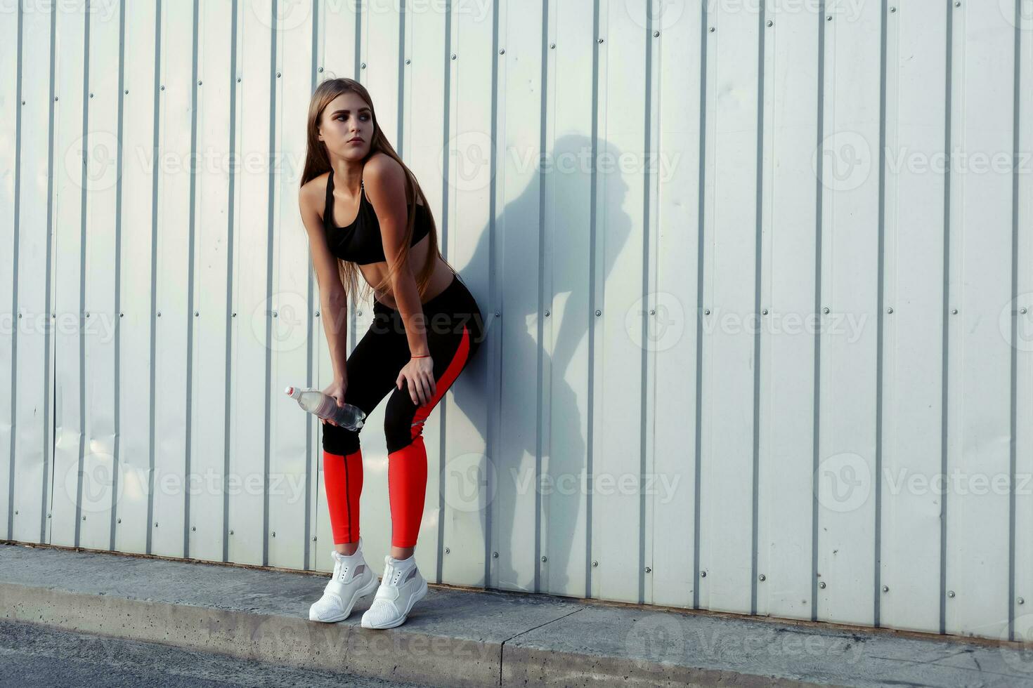 Female athlete drinking water while standing by a grey wall. photo