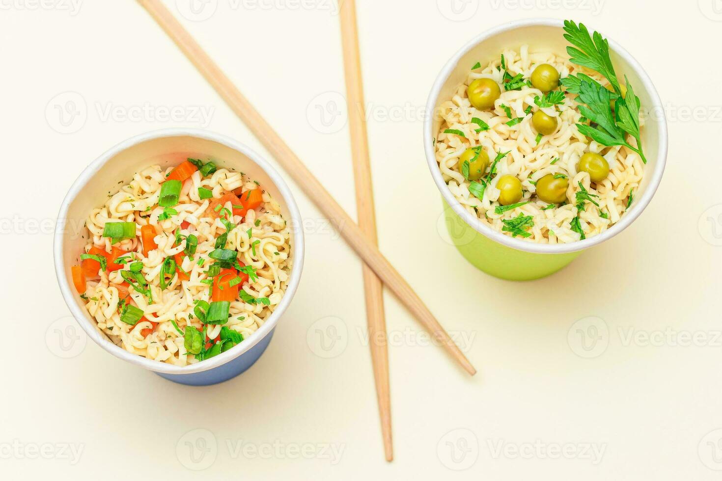 Prepared Instant Noodles with Green Pea, Carrot and Greens in Blue and Green Cups with Chopsticks on White Background. Quick Lunch or Unhealthy Fast Food photo