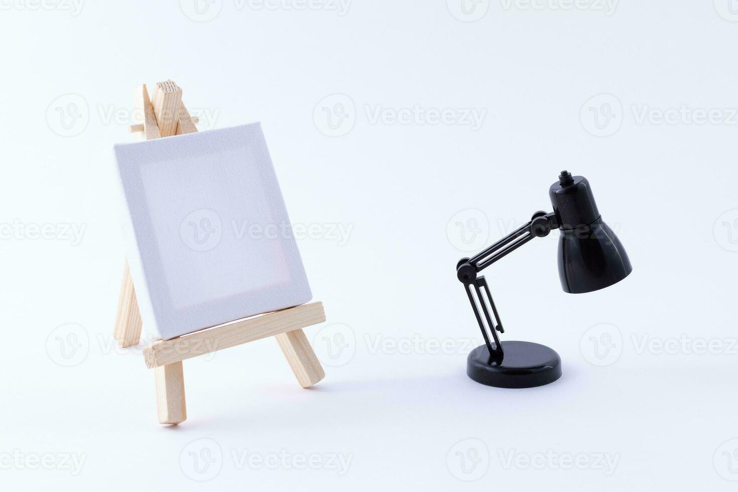 Wooden Easel Miniature with Blank White Square Canvas for Artists and Painters Mockup. Mini Wooden Stand with Clean Artboard and Small Black Table Lamp on White Background, Copy Space photo