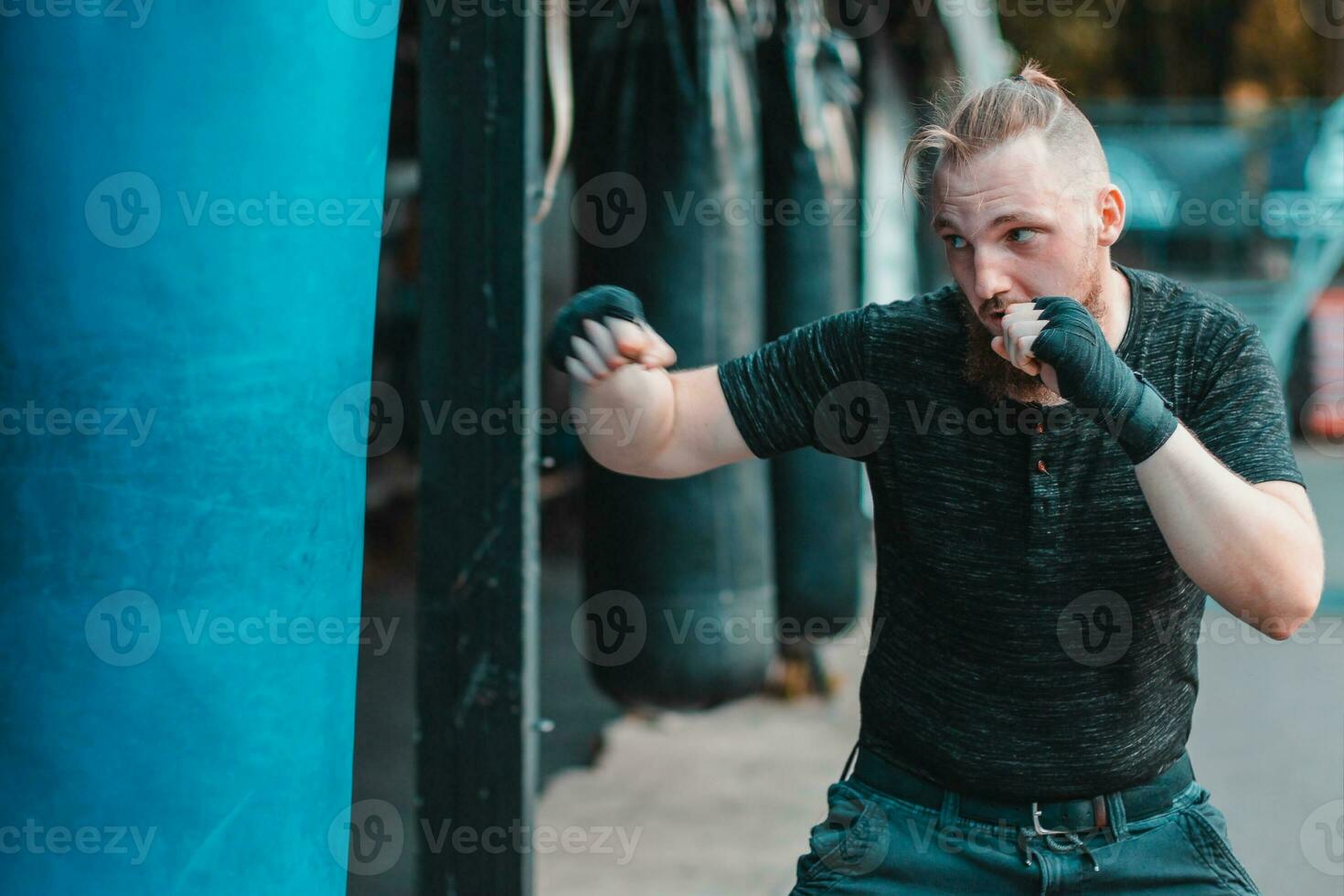 Street Fighter in Black Clothes and Bandages on the Wrist Boxing in Punching Bag Outdoors. Young Man Doing Box Training and Practicing His Punches at the Outside Gym photo