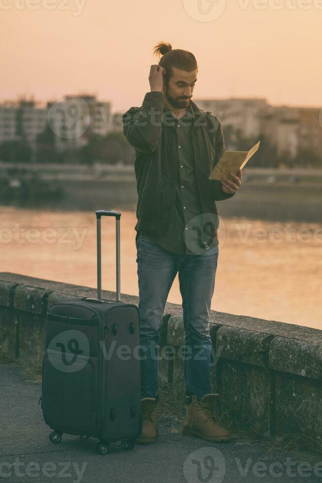 Handsome businessman with suitcase holding map of the city and standing by the river.Toned image photo