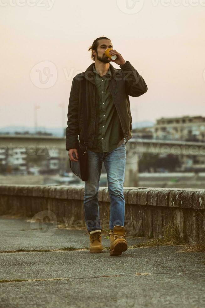 Handsome modern businessman with laptop bag drinking coffee and walking by the river.Toned image photo