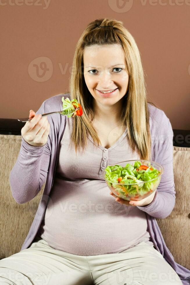 embarazada mujer sentado en el sofá y comiendo ensalada foto