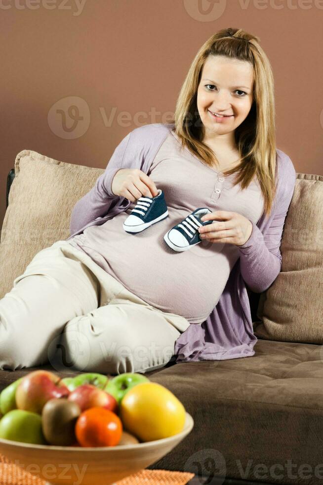 Smiling pregnant woman sitting on sofa and holding baby booties photo