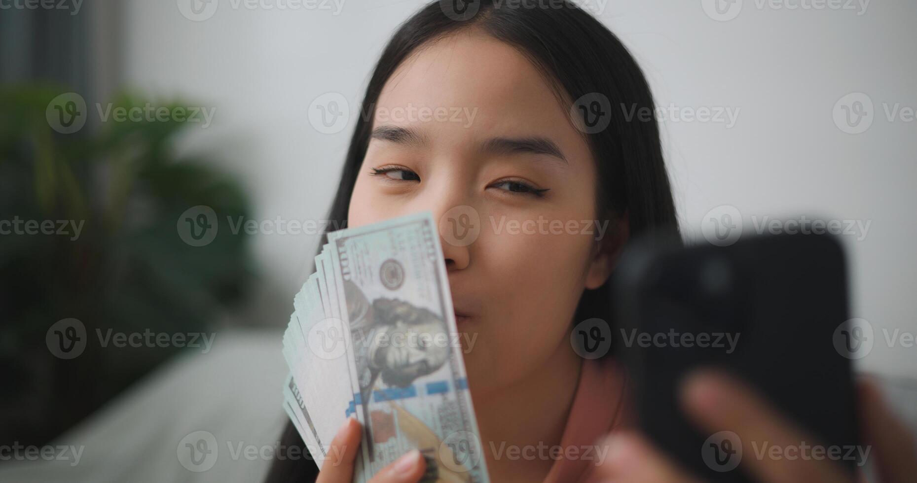 retrato de contento joven asiático mujer sentado en un emocionado estado, participación un en línea teléfono inteligente y dólar cuentas en su mano, ahorra dinero foto