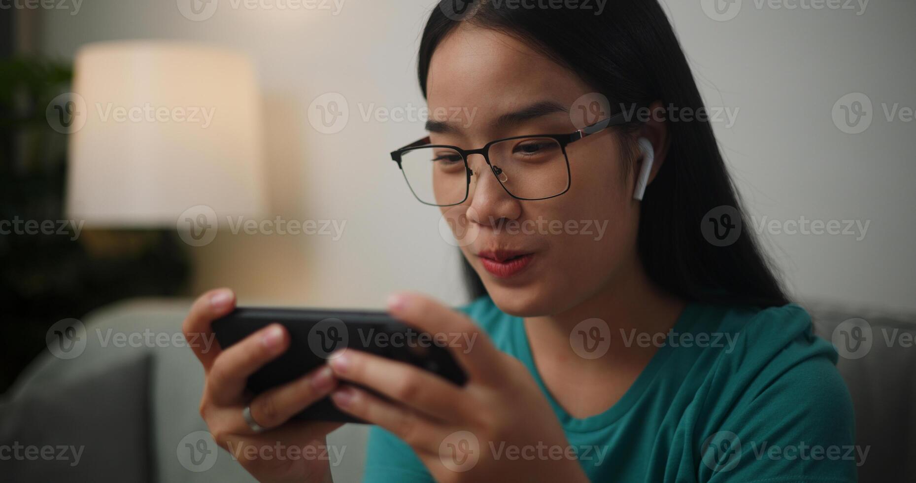 Portrait of Young Asian woman wearing glasses and headphones enjoys playing online esport games on smartphone sitting on sofa in the living room at home,Gamer lifestyle concept. photo