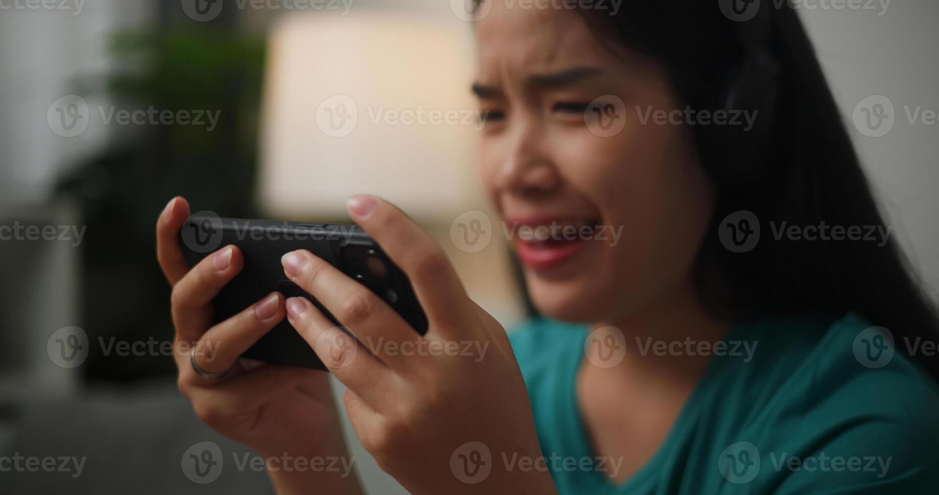 Portrait close up of Young Asian woman wearing glasses and headphones enjoys playing online esport games on smartphone sitting on sofa in the living room at home,Gamer lifestyle concept. photo