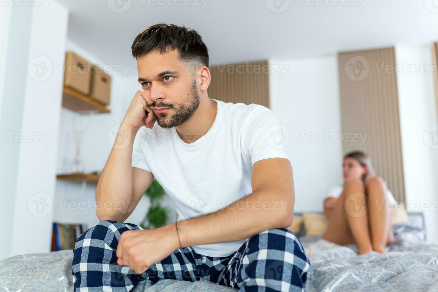 Sad thoughtful man after arguing with girlfriend.Relationship difficulties, conflict and family concept,unhappy couple having problems at bedroom. Sad guy sitting on bed,girlfriend in the background photo
