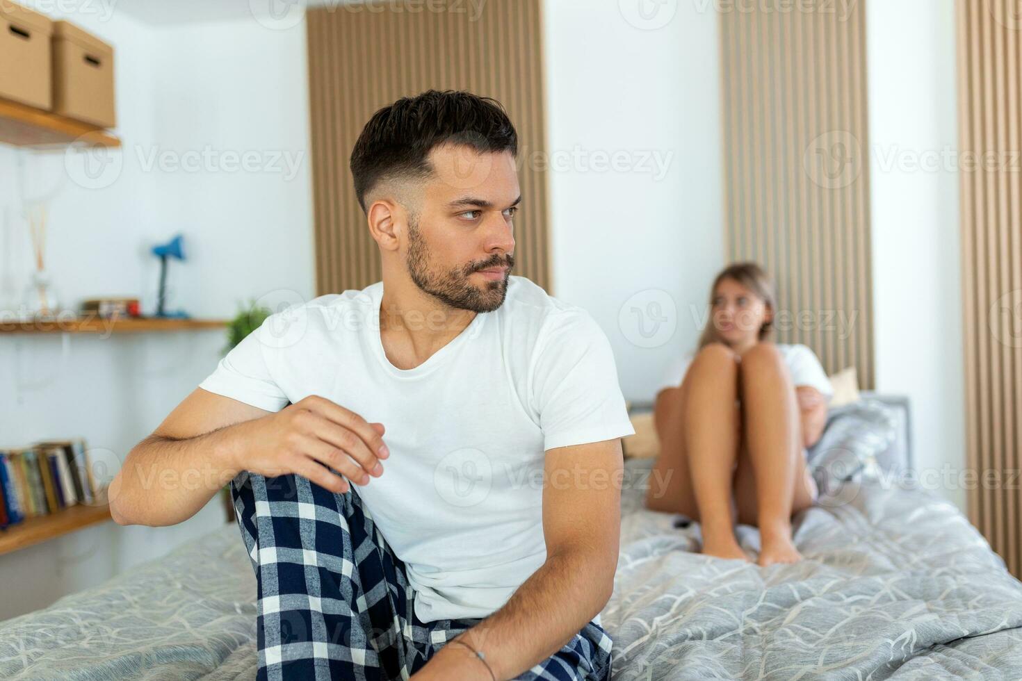 Sad thoughtful man after arguing with girlfriend.Relationship difficulties, conflict and family concept,unhappy couple having problems at bedroom. Sad guy sitting on bed,girlfriend in the background photo