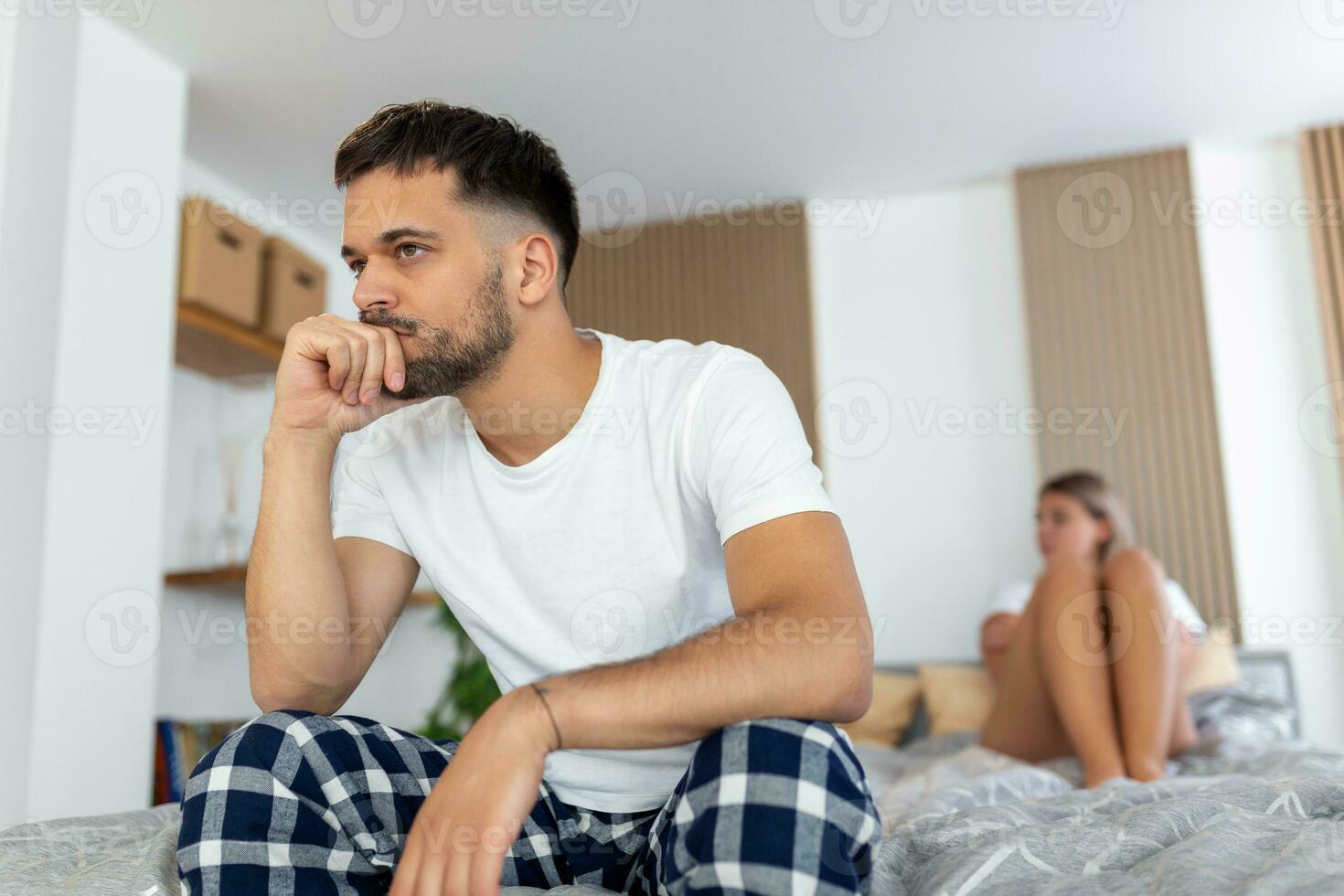 Sad thoughtful man after arguing with girlfriend.Relationship difficulties, conflict and family concept,unhappy couple having problems at bedroom. Sad guy sitting on bed,girlfriend in the background photo