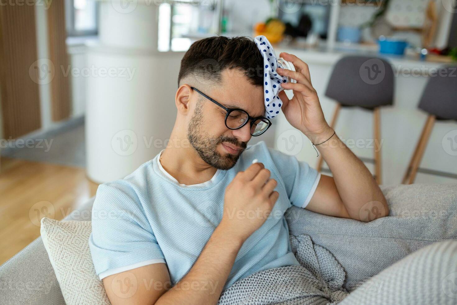 Man feeling bad, sitting on the sofa with headache and applying ice bag photo