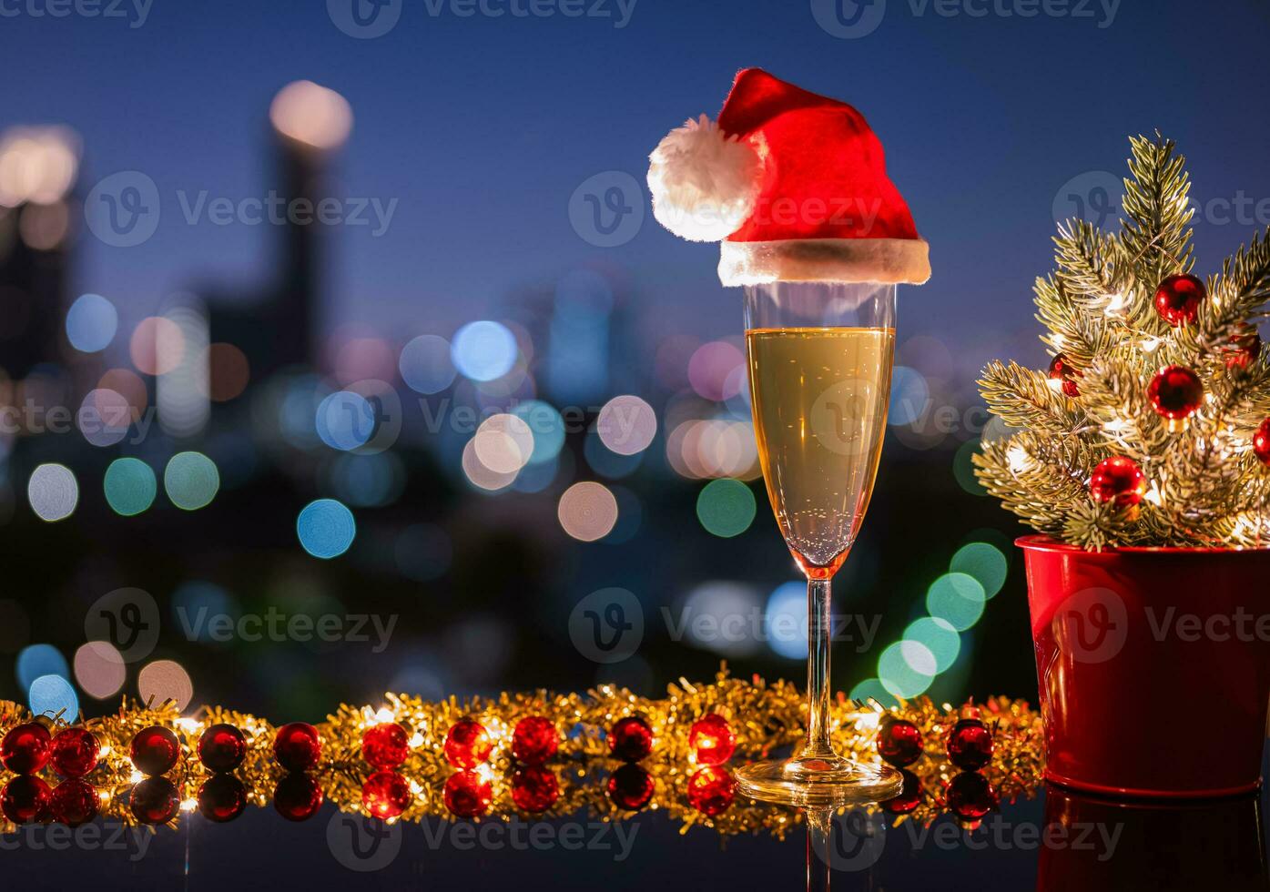A glass of white wine that have santa claus hat on it with Christmas ornament decoration put on table with city bokeh light background. Christmas dining concept. photo