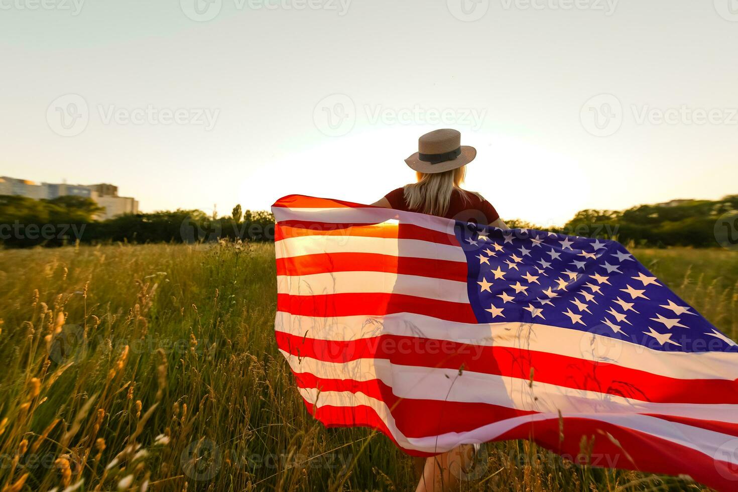 Beautiful young girl holding an American flag in the wind in a field of rye. Summer landscape against the blue sky. Horizontal orientation. photo