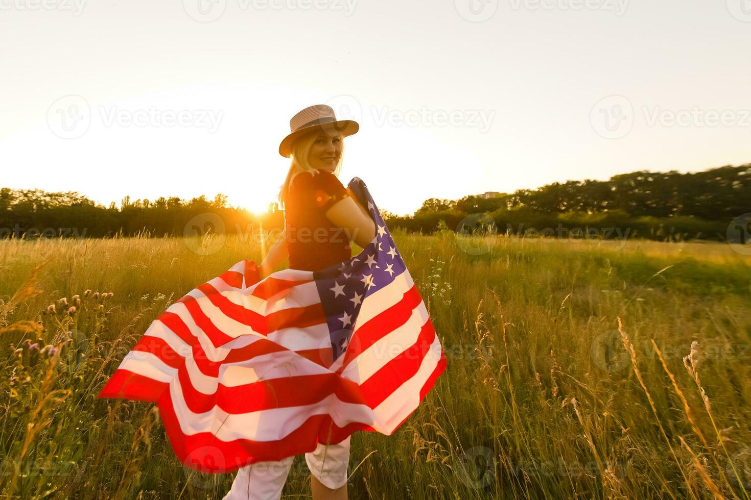 Beautiful young girl holding an American flag in the wind in a field of rye. Summer landscape against the blue sky. Horizontal orientation. photo