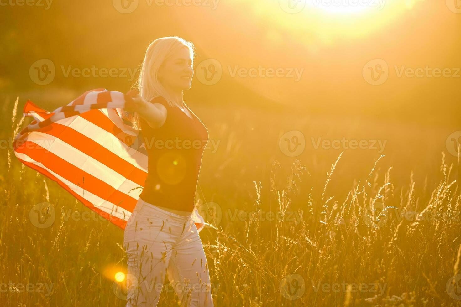 Beautiful young girl holding an American flag in the wind in a field of rye. Summer landscape against the blue sky. Horizontal orientation. photo