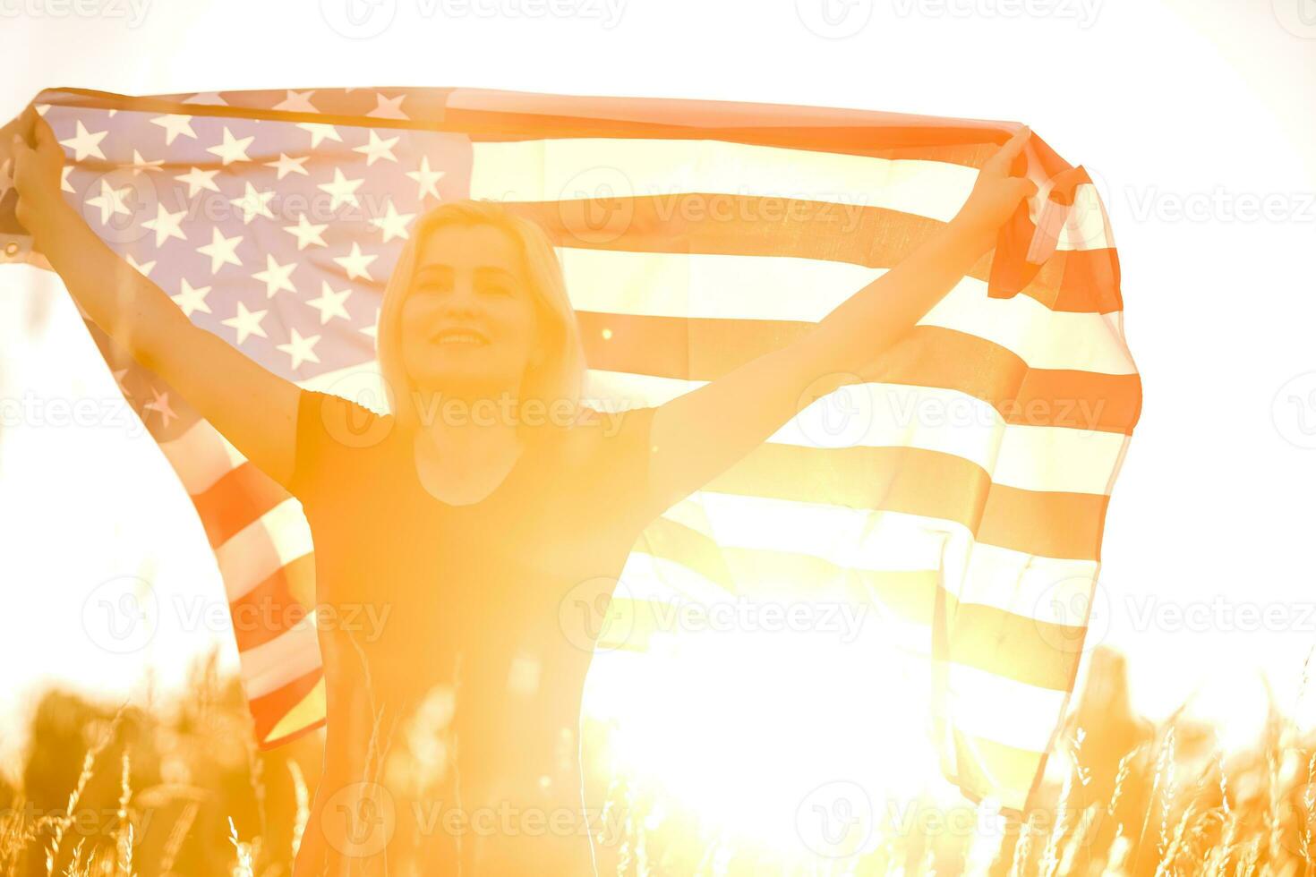 Beautiful young girl holding an American flag in the wind in a field of rye. Summer landscape against the blue sky. Horizontal orientation. photo