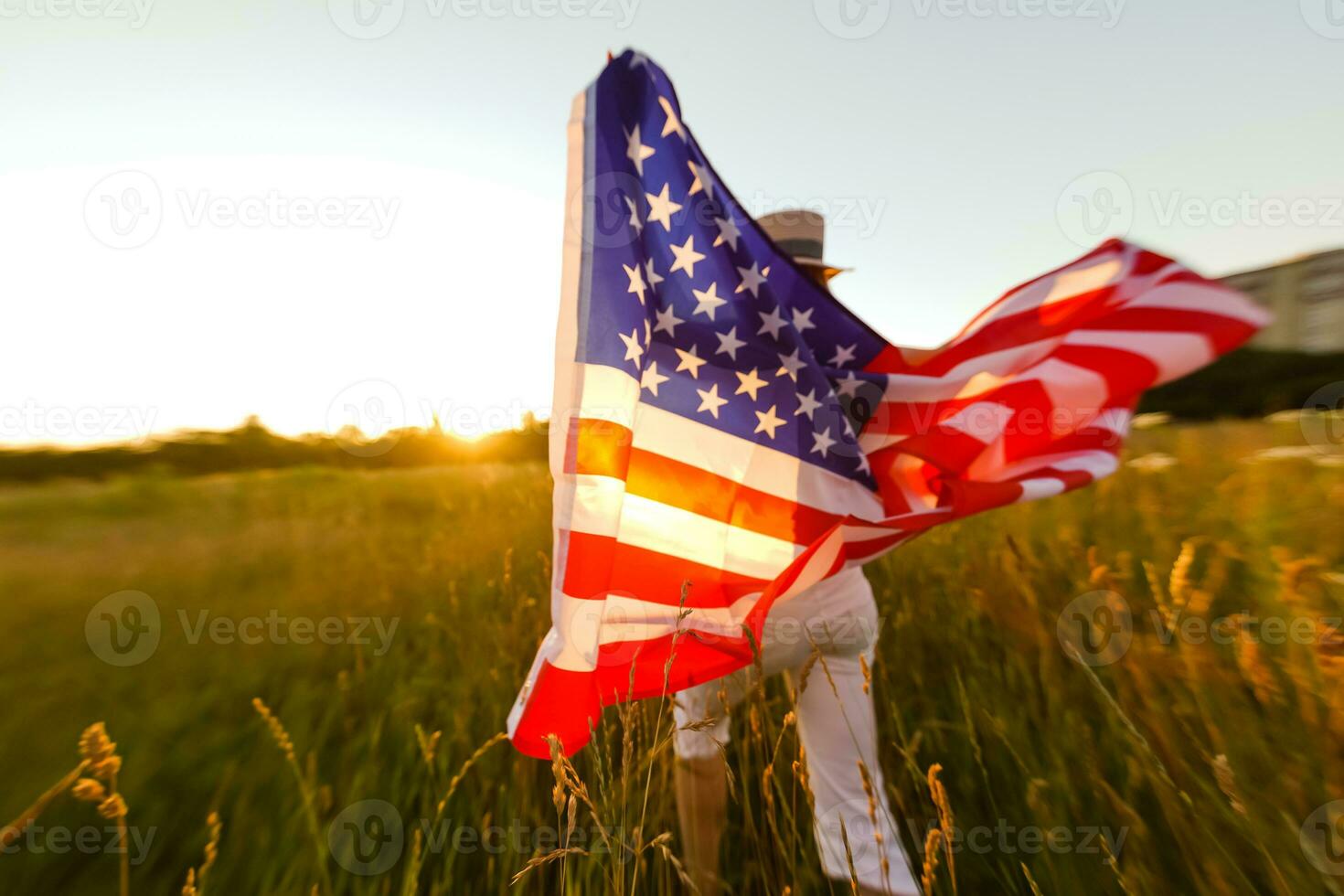 Beautiful young girl holding an American flag in the wind in a field of rye. Summer landscape against the blue sky. Horizontal orientation. photo