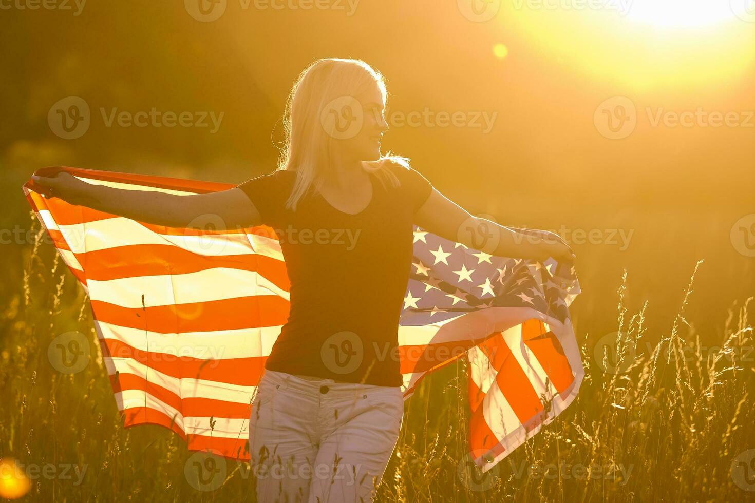 Beautiful young girl holding an American flag in the wind in a field of rye. Summer landscape against the blue sky. Horizontal orientation. photo