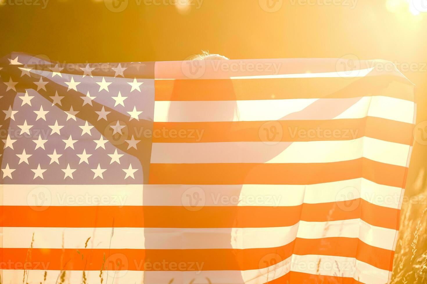 Beautiful young girl holding an American flag in the wind in a field of rye. Summer landscape against the blue sky. Horizontal orientation. photo