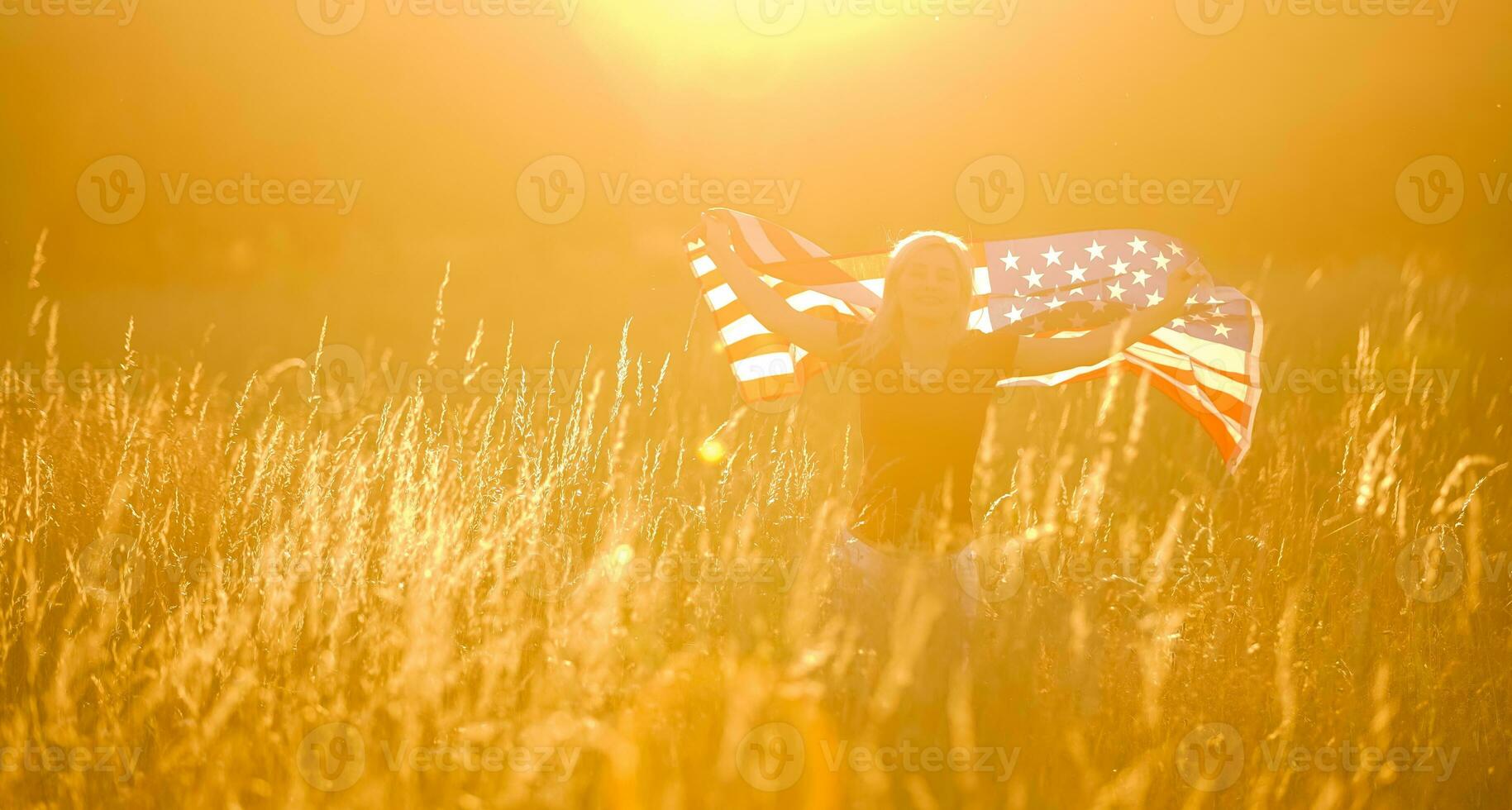 Beautiful young girl holding an American flag in the wind in a field of rye. Summer landscape against the blue sky. Horizontal orientation. photo