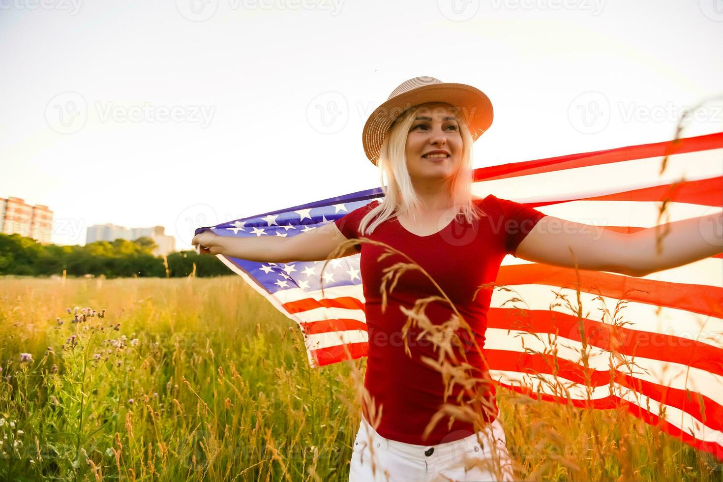 Beautiful young girl holding an American flag in the wind in a field of rye. Summer landscape against the blue sky. Horizontal orientation. photo