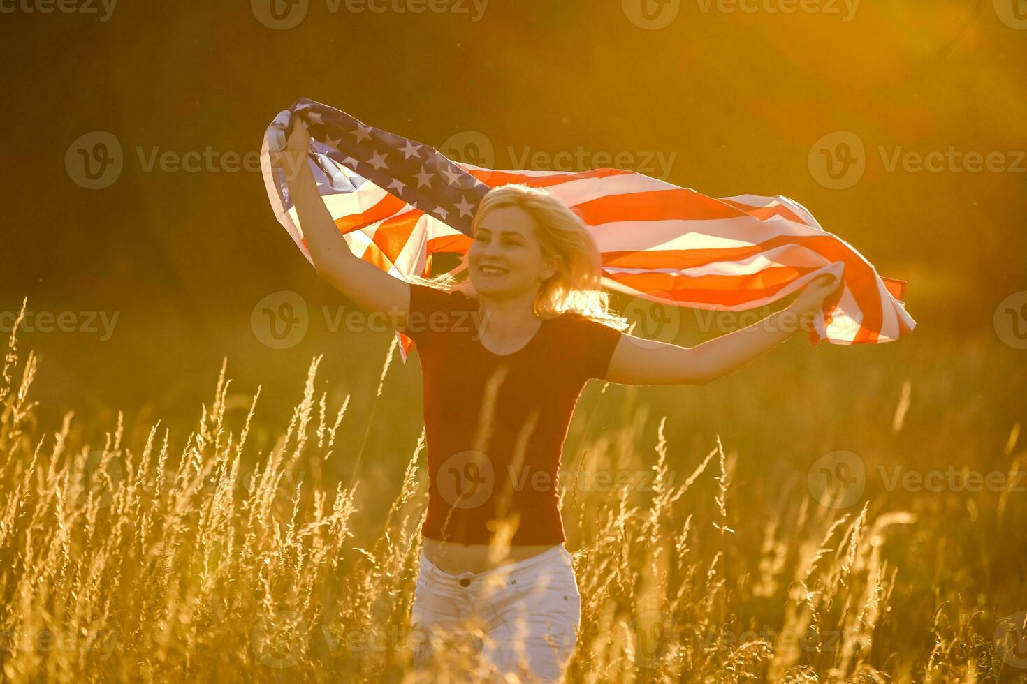 Beautiful young girl holding an American flag in the wind in a field of rye. Summer landscape against the blue sky. Horizontal orientation. photo