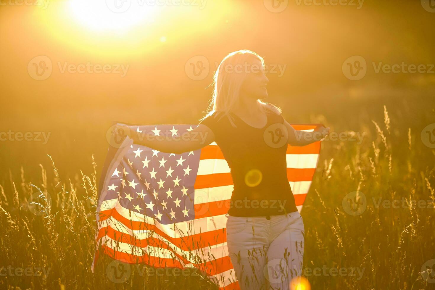 Young happy girl running and jumping carefree with open arms over wheat field. Holding USA flag. Toned image. Selective focus. photo
