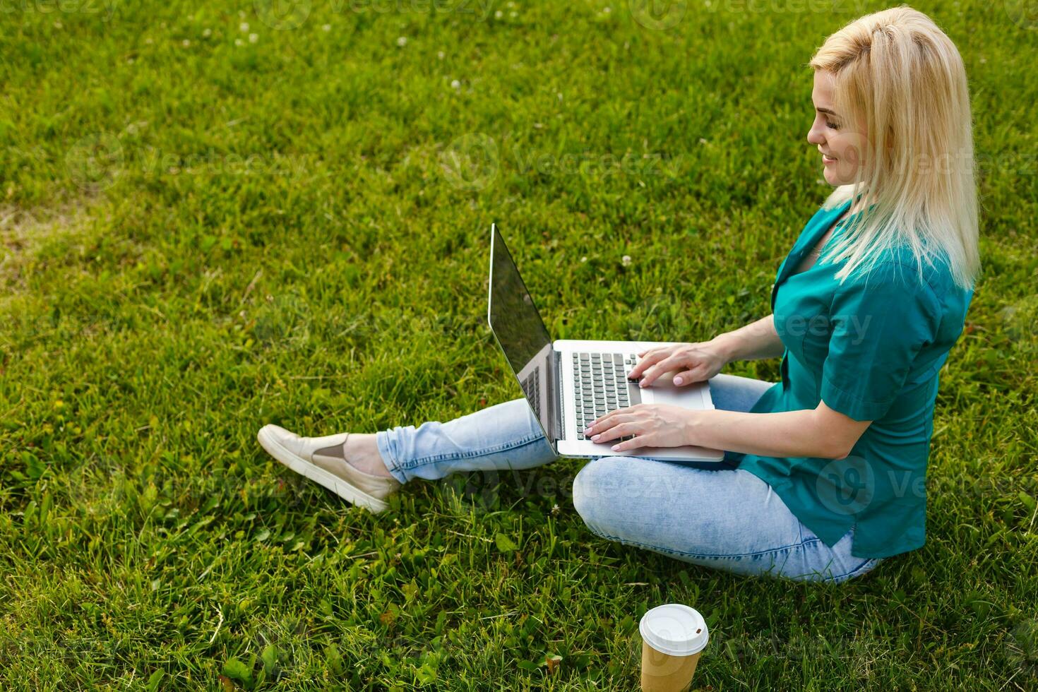 Top view of woman sitting in park on the green grass with laptop, hands on keyboard. Computer screen mockup. Student studying outdoors. Copy space for text photo