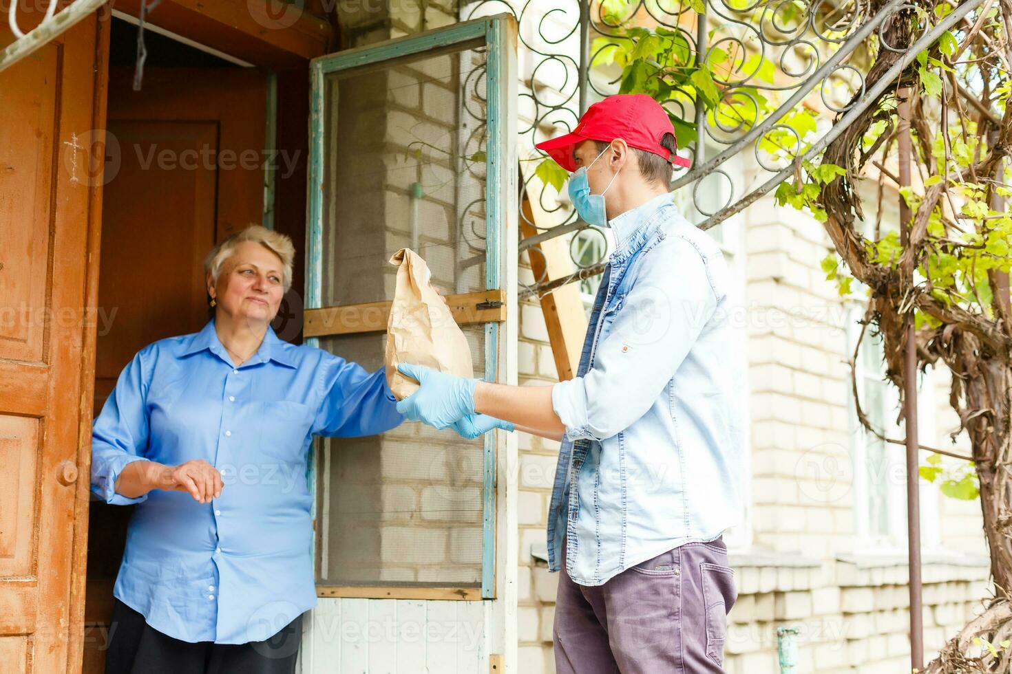 Young male volunteer in mask gives an elderly woman boxes with food near her house. Son man helps a single elderly mother. Family support, caring. Quarantined, isolated. Coronavirus covid-19. Donation photo