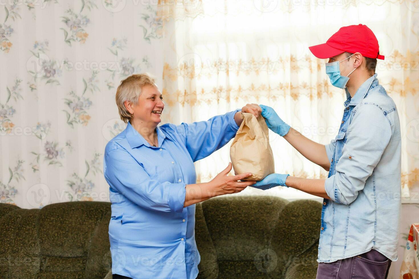 Young male volunteer in mask gives an elderly woman boxes with food near her house. Son man helps a single elderly mother. Family support, caring. Quarantined, isolated. Coronavirus covid-19. Donation photo