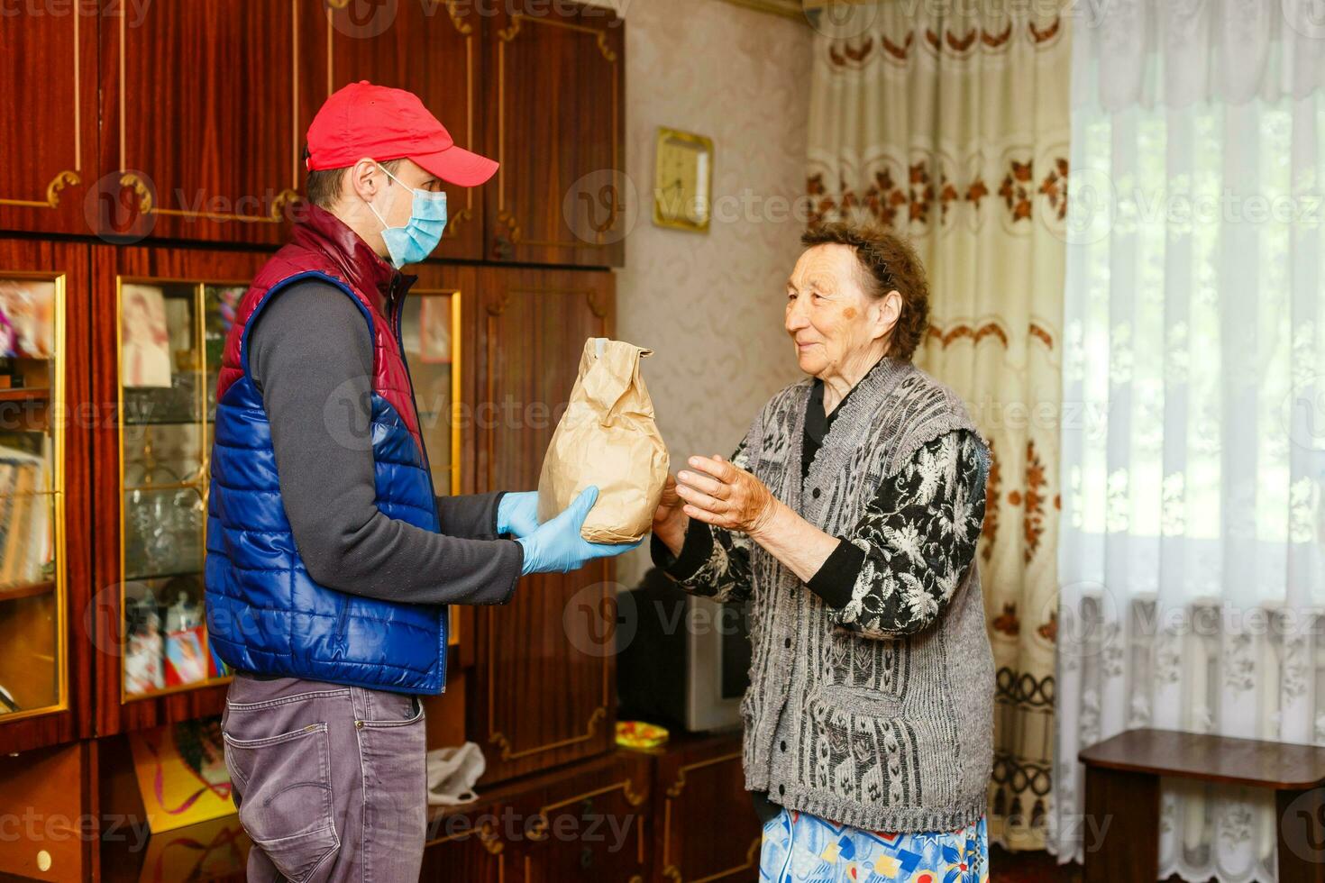 Young male volunteer in mask gives an elderly woman boxes with food near her house. Son man helps a single elderly mother. Family support, caring. Quarantined, isolated. Coronavirus covid-19. Donation photo