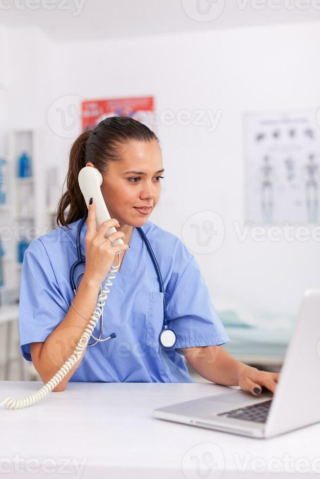 Medical practitioner having a conversation with patient on phone from hospital office and checking appointment. Health care physician sitting at desk using computer in modern clinic looking at monitor. photo