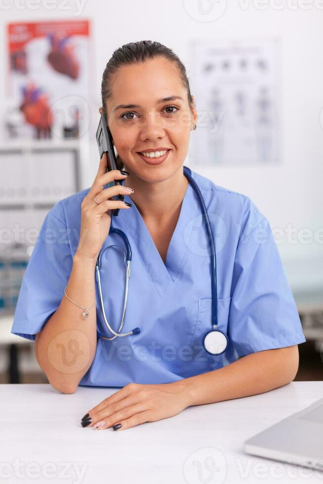 Medical staff talking with patient on phone from hospital office about diagnosis smiling at camera. Female nurse, doctor having a phone conversation with sick person during consultation, medicine. photo