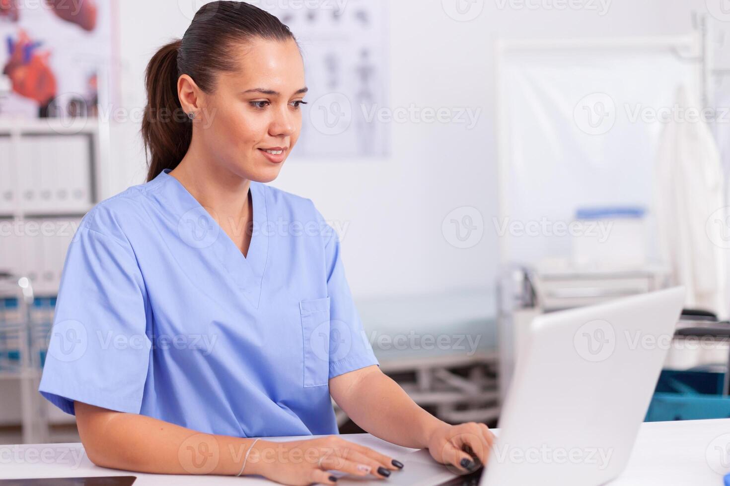 Medical physician typing on laptop sitting at desk wearing blue uniform. Health care physician using computer in modern clinic looking at monitor, medicine, profession, scrubs. photo