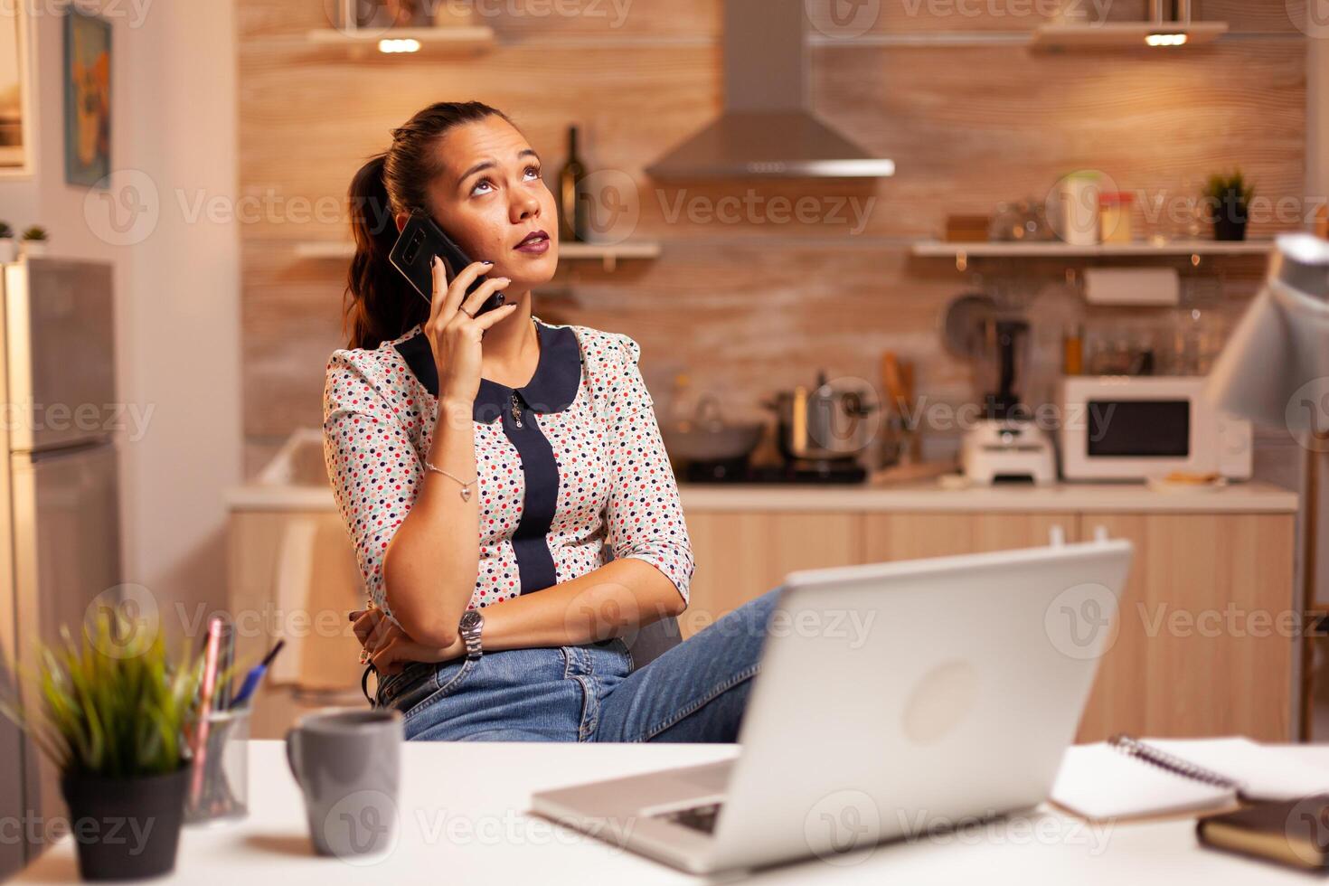 Freelance woman having a phone coversation with a client late at night from home kitchen. Employee using modern technology at midnight doing overtime for job, business, career, network, lifestyle. photo