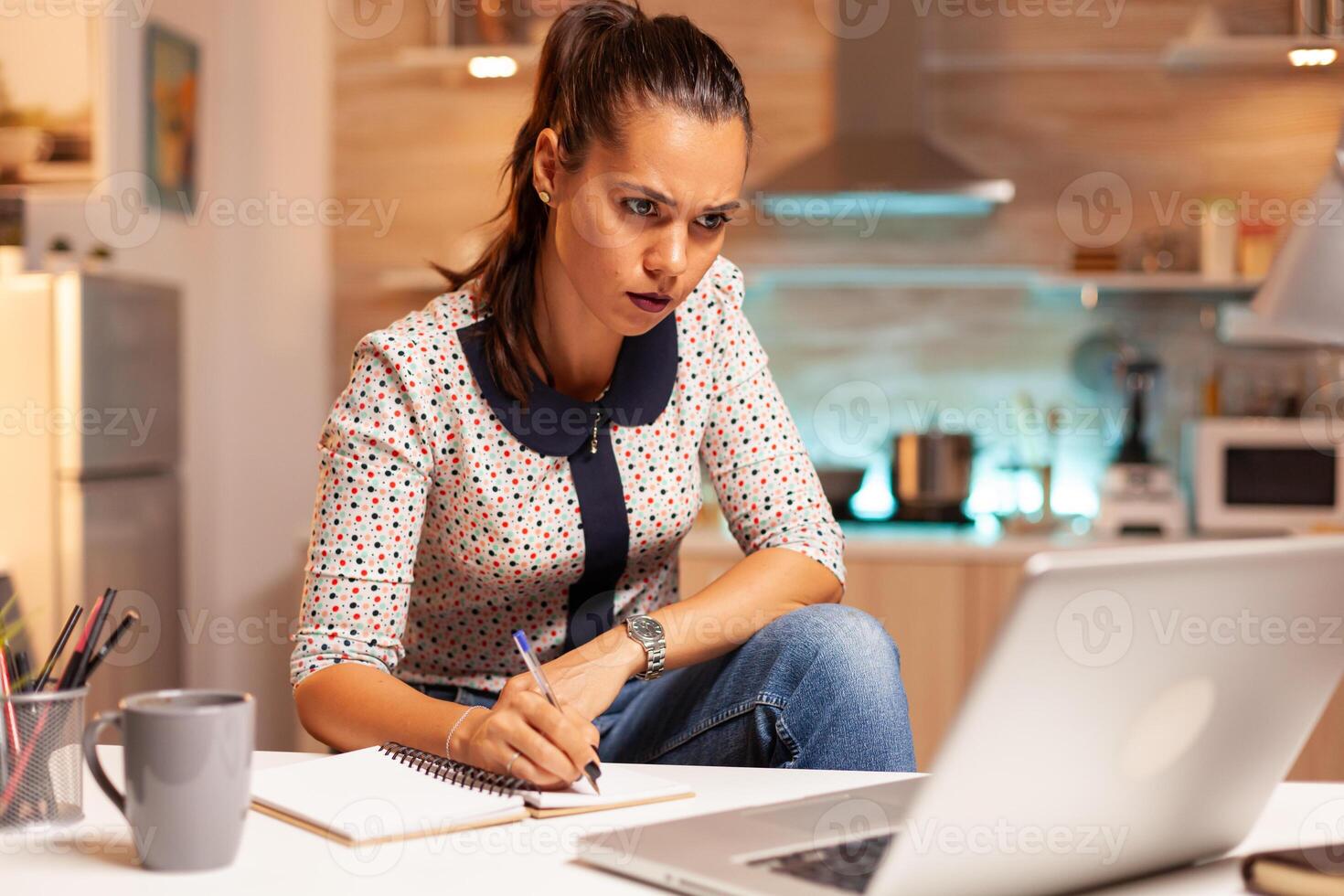 Businesswoman writing notes while working late on computer in home kitchen. Employee using modern technology at midnight doing overtime for job, business, busy, career, network, lifestyle ,wireless. photo