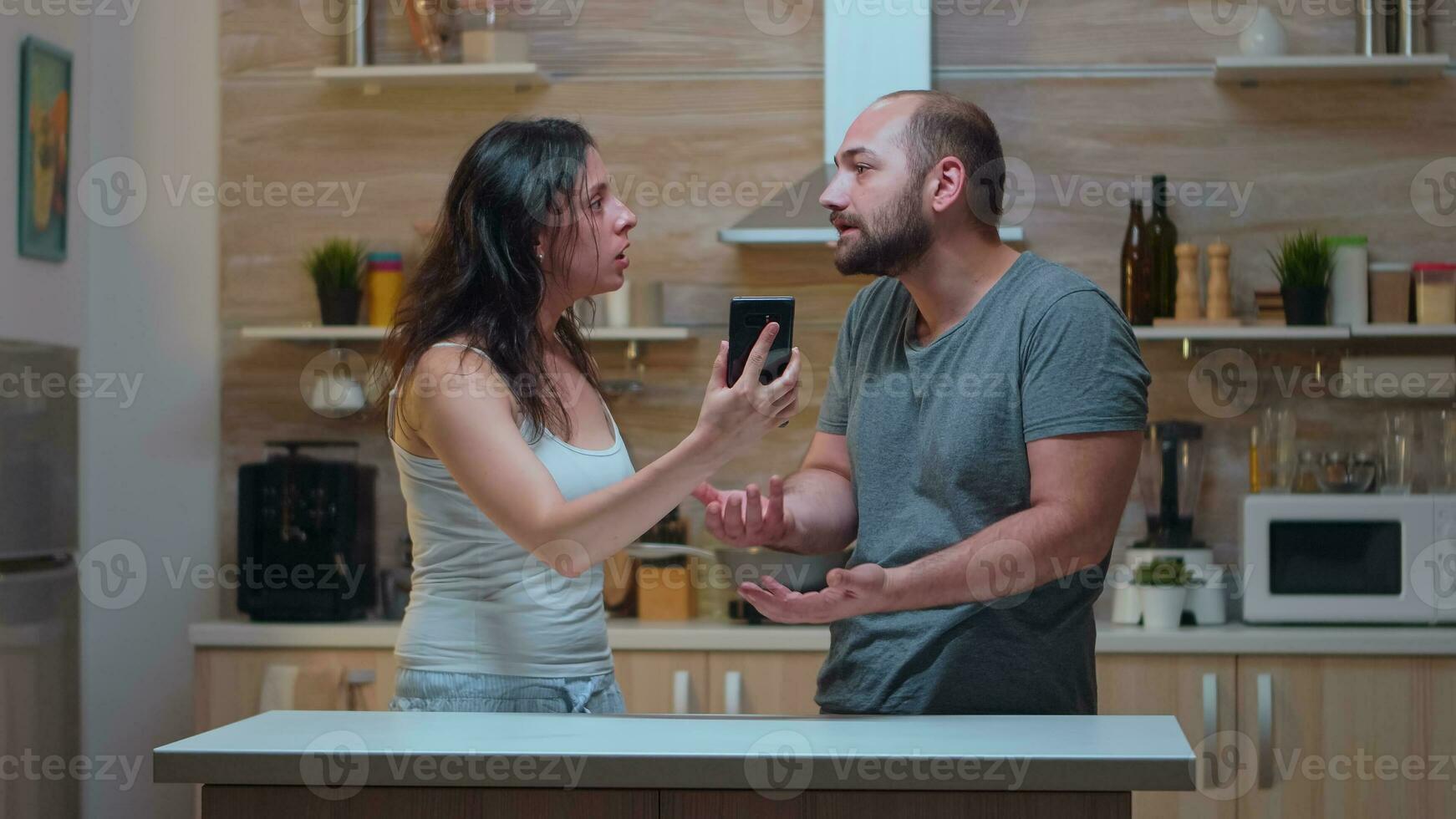 Wife screaming at unfaithful husband in the kitchen. Jealous woman cheated angry frustrated offended irritated accusing her man of infidelity showing him messages on smartphone yelling desperate. photo