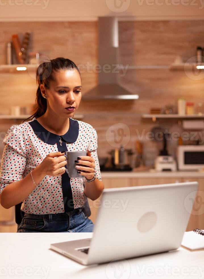 Woman sitting in kitchen lat at night working on a project for work using laptop and holding cup of coffee. Employee using modern technology at midnight doing overtime for job, business, busy, career. photo