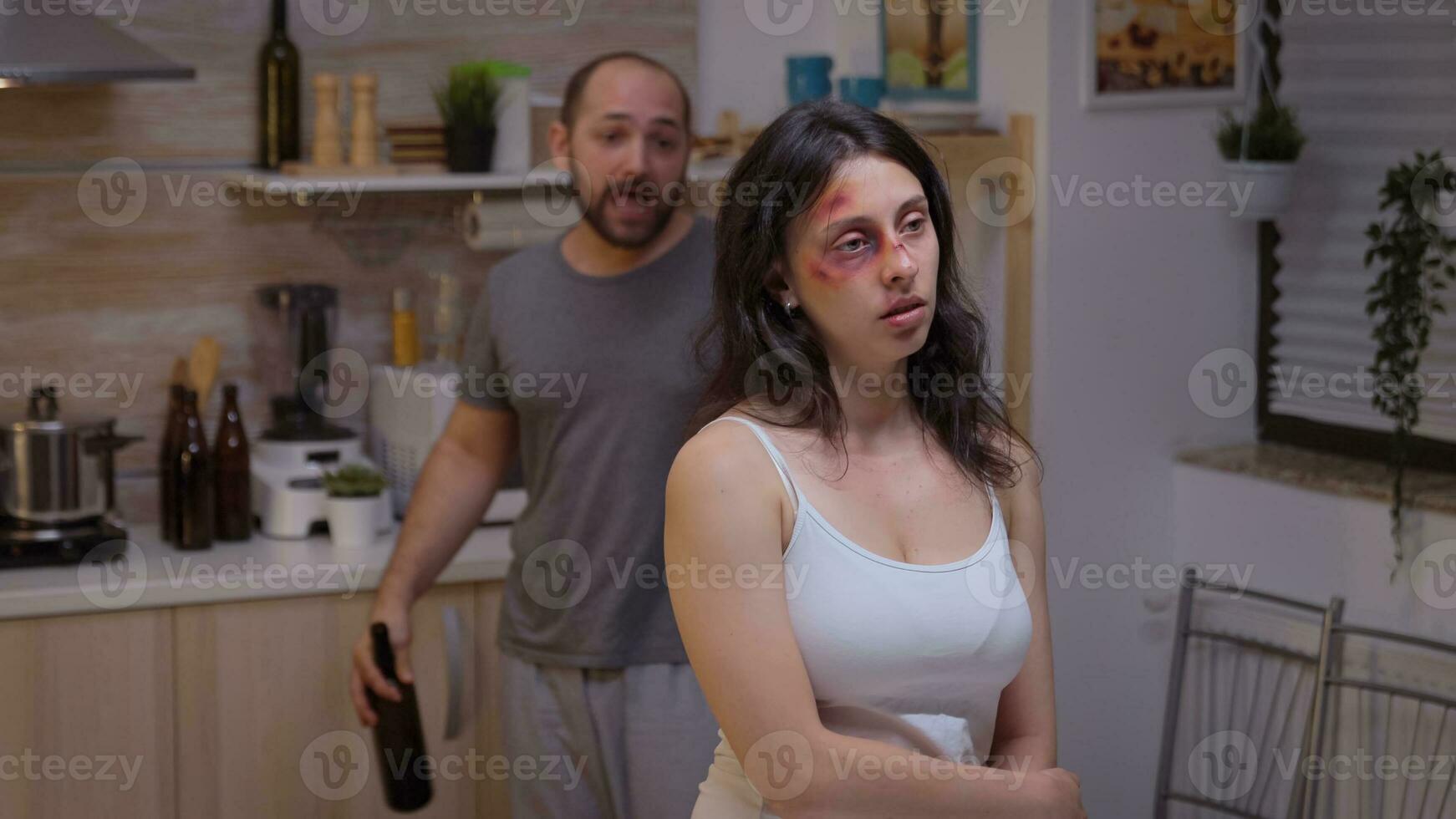 Alcoholic man screaming at beaten woman walking drunk through the kitchen. Traumatised abused terrified beaten wife covered in bruises suffering injury from violent brutal man. photo