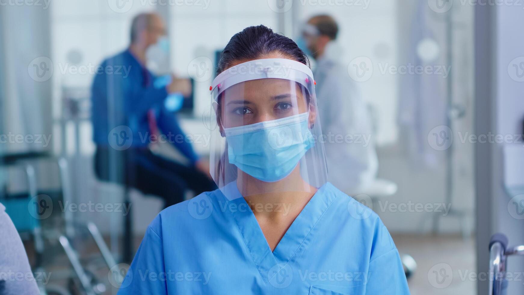 Medical assistant with visor and face mask against coronavirus looking at camera in hospital waiting area. Doctor consulting senior man in examination room. photo