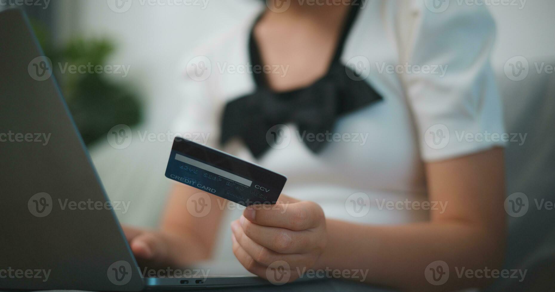 retrato de joven asiático mujer sentado en sofá en vivo habitación, hace en línea bancario pagos mediante el Internet desde banco tarjeta en portátil, compras en línea con crédito tarjeta. foto
