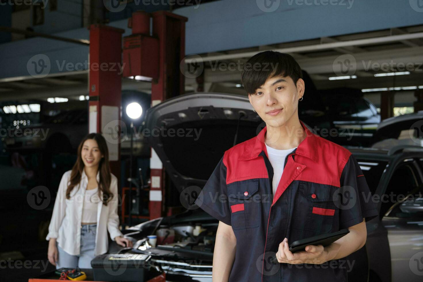 Portrait of young Asian male automotive mechanic worker with female customer's EV car at maintenance garage, happy smiles, professional service station, check and repair in auto transport industry. photo