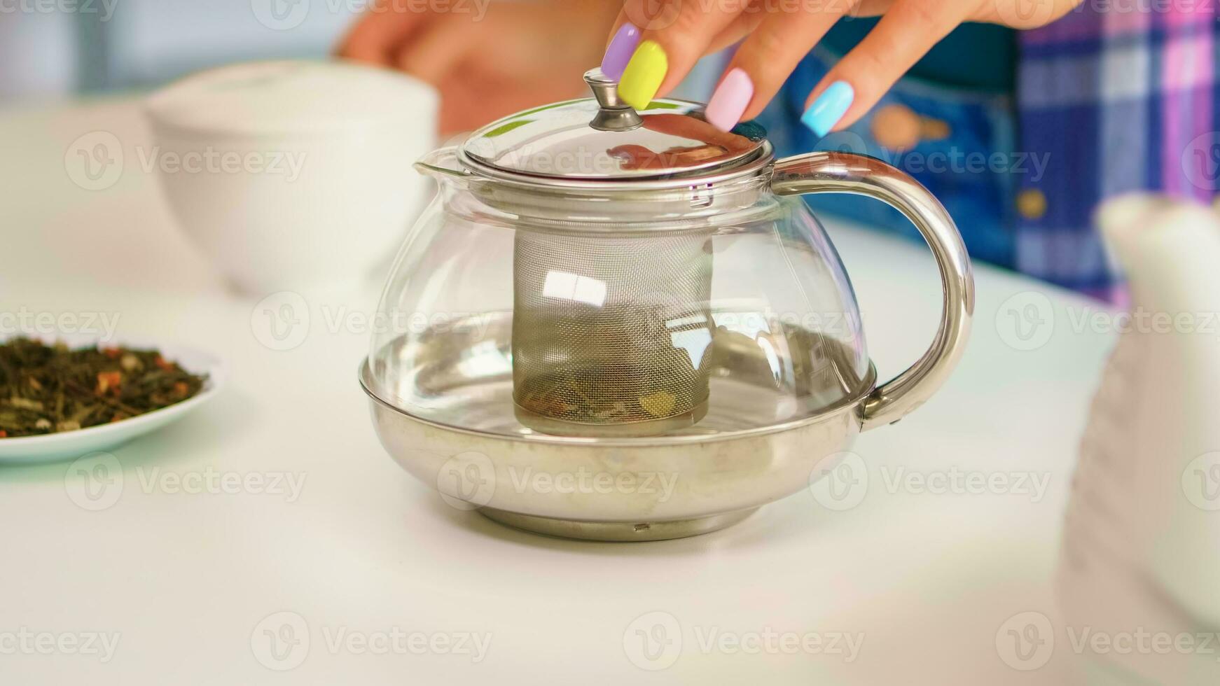 Close up of a woman brewing green tea in the morning, for breakfast, in a modern kitchen using teapot sitting near the table. SHe is putting with hands, pressing herbal, healthy, tea leaves, in pot photo