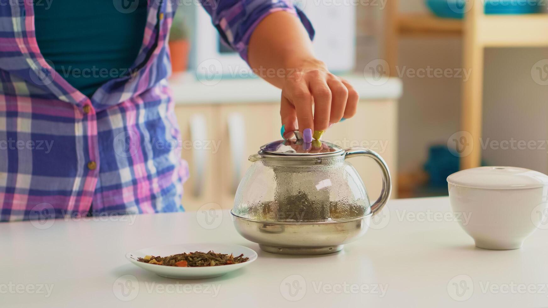 Close up of young woman adding hot water over herbal, tea leaves. Housewife preparing green tea for breakfast using teapot in a modern kitchen sitting near the table holding kettle waiting to infuse. photo