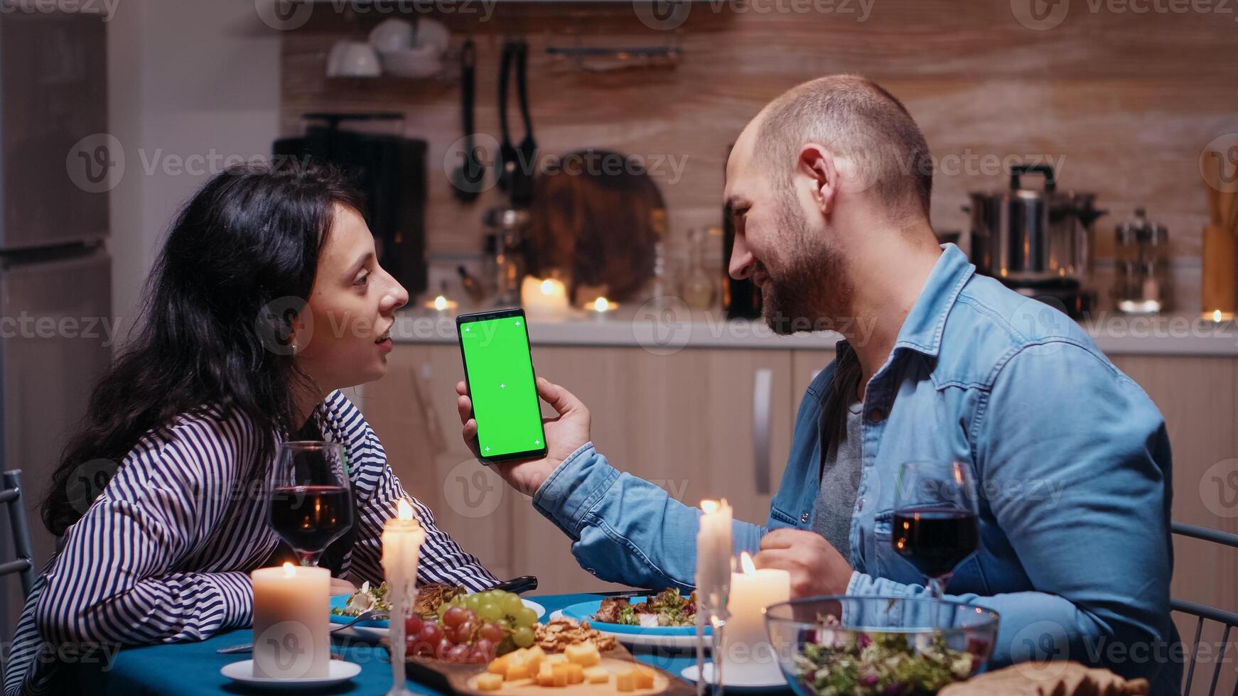 Young happy couple holding green screen phone at dinner. Cheerful people looking at mockup template chroma key isolated smart phone display using techology internet sitting at the table in kitchen. photo