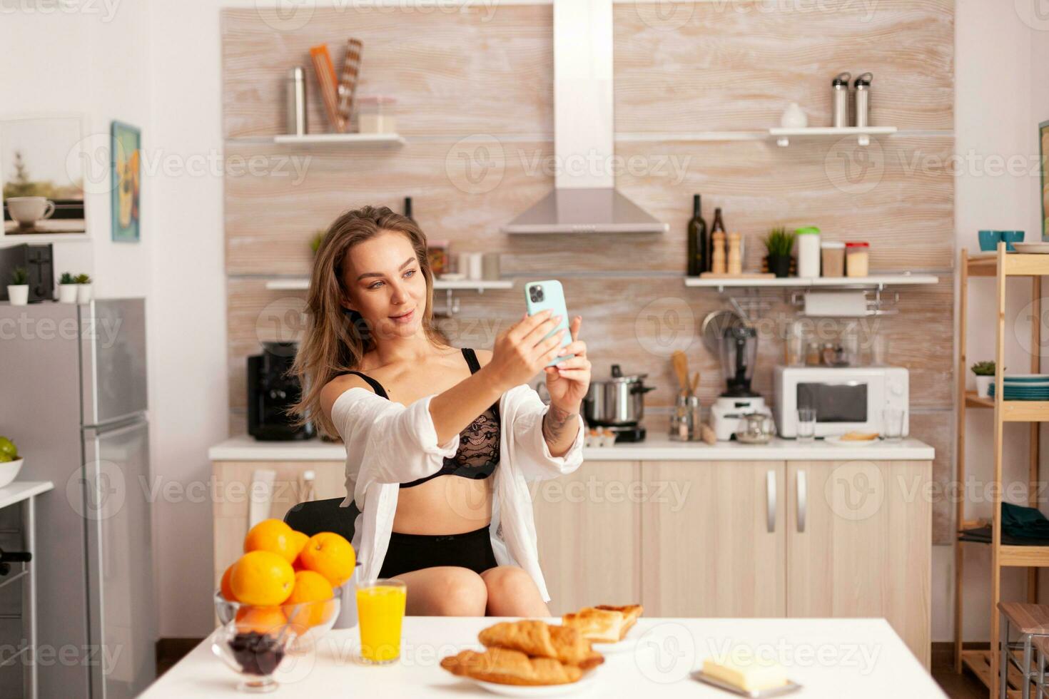 Seductive woman in sexy lingerie taking selfie using smartphone in home kitchen. Attractive lady with tattoos using smartphone wearing temping underwear in the morning. photo