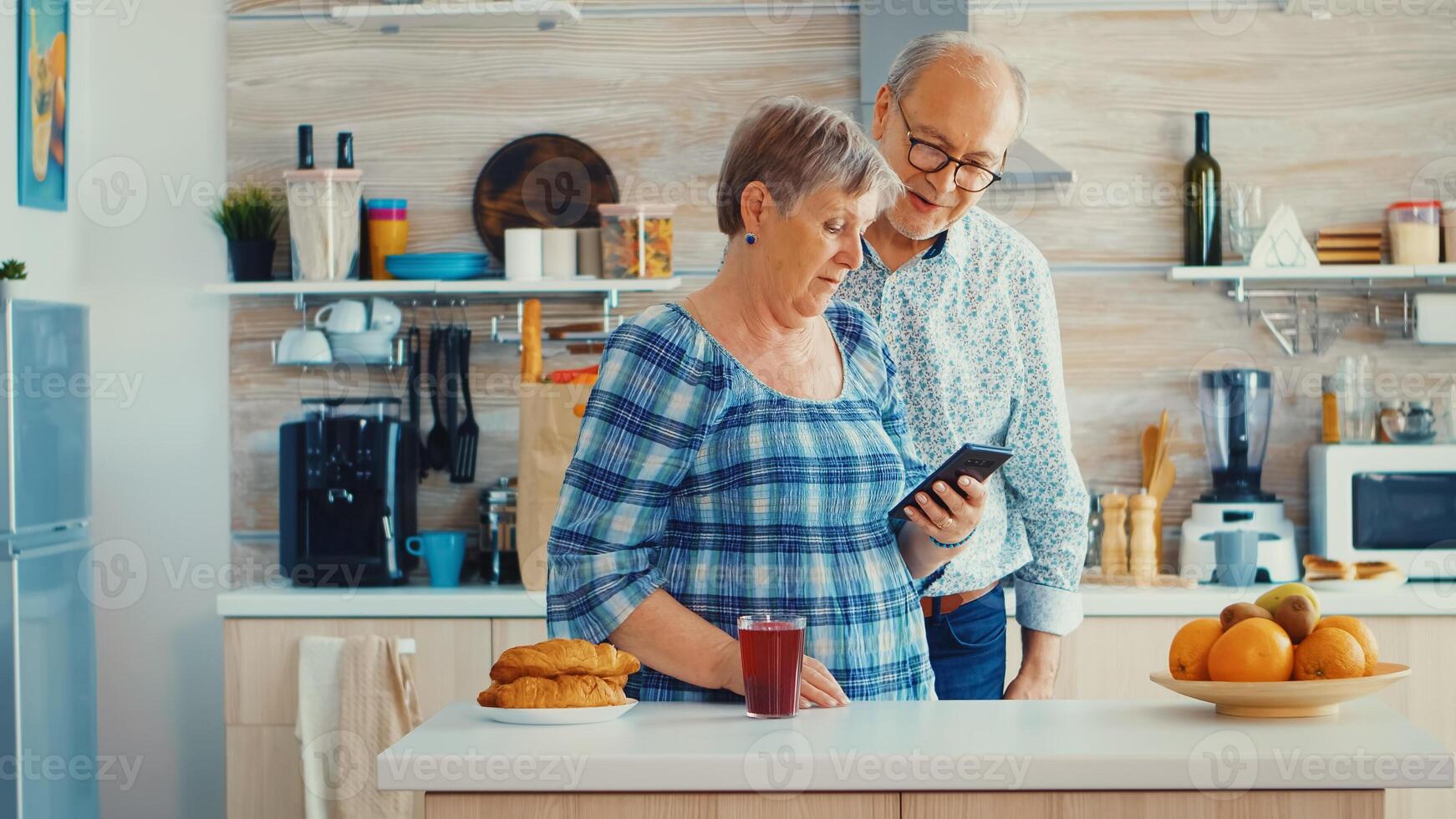 Older couple during video chat with family using smartphone in kitchen. Grandparents online conversation. Elderly persons with modern technology in retirement age using mobile apps photo