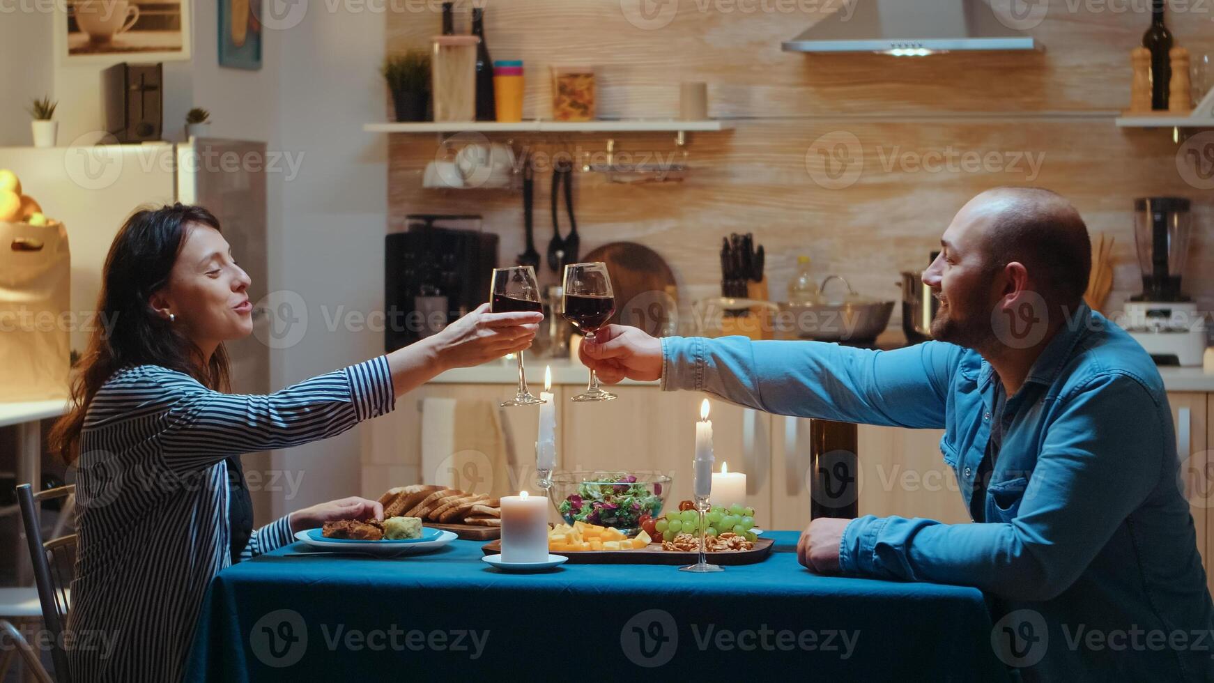 Loving young couple raising red wine glass and toasting while enjoying romantic dinner at home in the kitchen. Happy in love people eating meal, celebrating anniversary in dining room, romantic toast photo