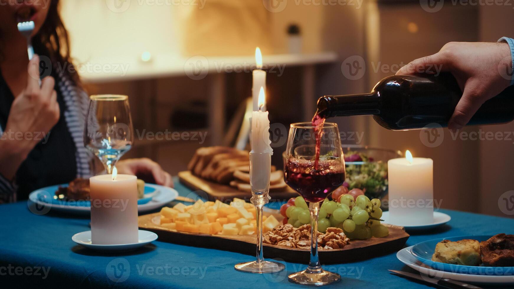 Close up of young man pouring red wine in wife glass. Romantic caucasian happy couple sitting at the table in kitchen celebrating with candle lights, love and anniversary. Romance surprise in relation photo